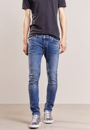 Personne portant un jean slim bleu, un t-shirt noir et des baskets montantes argentées, debout sur un sol et un fond neutres.