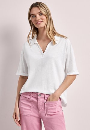 Femme aux cheveux blonds portant un t-shirt blanc à manches courtes avec col en V et un pantalon rose, debout avec une main dans la poche sur un fond clair.
