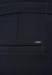 Dunkelblauer Stoff mit glatter Textur, ausgestattet mit einer Rückentasche und einem kleinen silberfarbenen Metalletikett mit der Aufschrift "CECIIL."