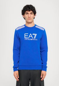 Modré mikina s dlhým rukávom a okrúhlym výstrihom. Na prednej strane sa nachádza biele logo EA7 a Emporio Armani. Horizontálne pruhy na ramenách.