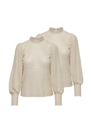 Frau mit langen Haaren, die einen beigen strukturierten Pullover, weite braune Hosen und braune spitze Pumps trägt und mit einer Hand nahe am Gesicht steht.