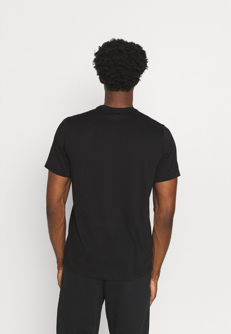 Homme aux cheveux courts et bouclés, de dos, portant un t-shirt noir uni à manches courtes et un pantalon noir sur fond blanc.