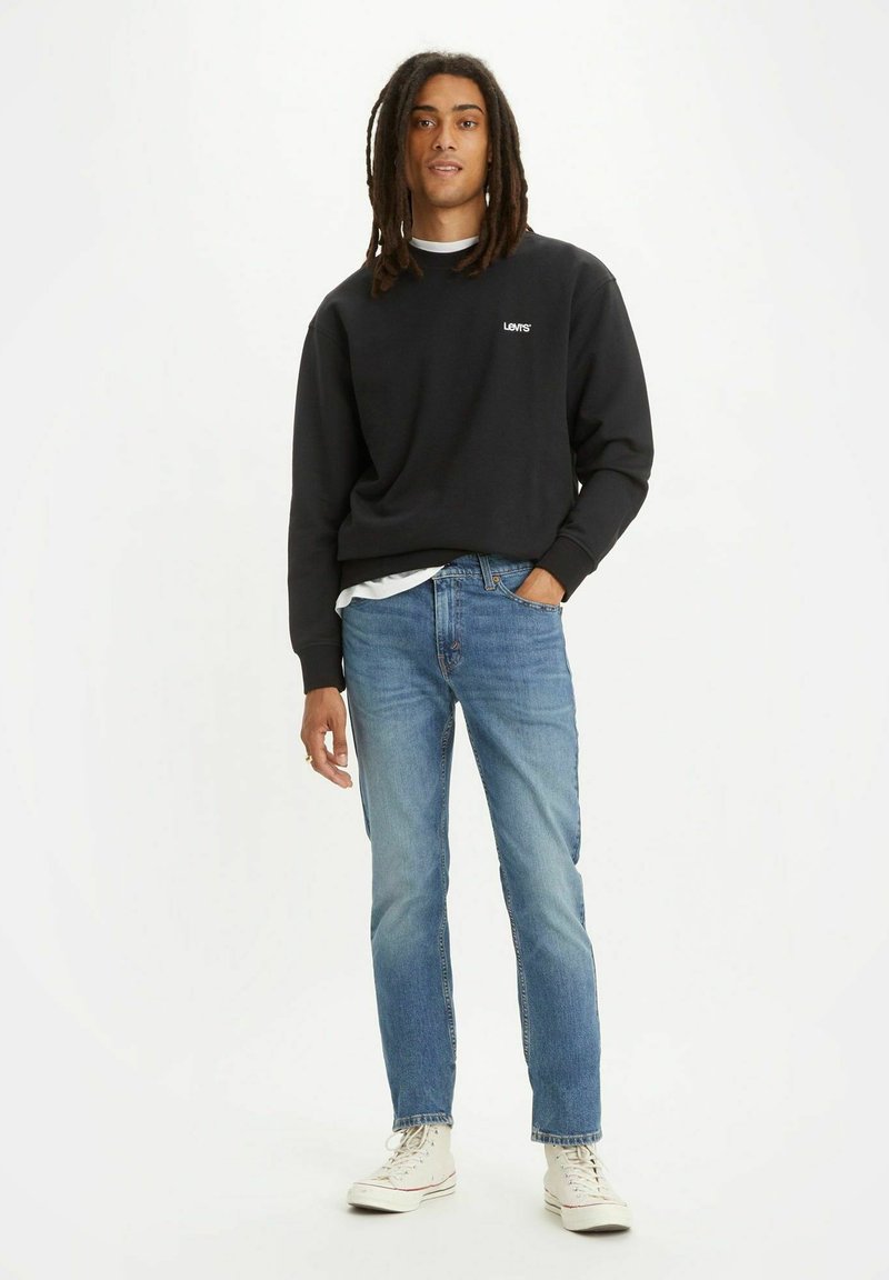Hombre con rastas que lleva una sudadera negra de Levi's, jeans azul claro y zapatillas altas blancas, de pie con una mano en el bolsillo.