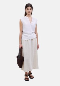 Top sin mangas de lino blanco con escote en forma de ojal, combinado con una larga falda de lino beige y un bolso de malla marrón oscuro. Sandalias negras.