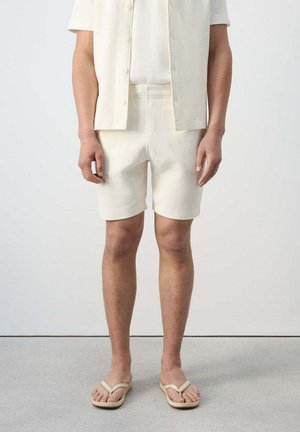 Modelo masculino vestido con pantalones cortos de punto color crema, camisa de manga corta a juego con botones y chanclas beige, de pie sobre un suelo gris con fondo blanco.