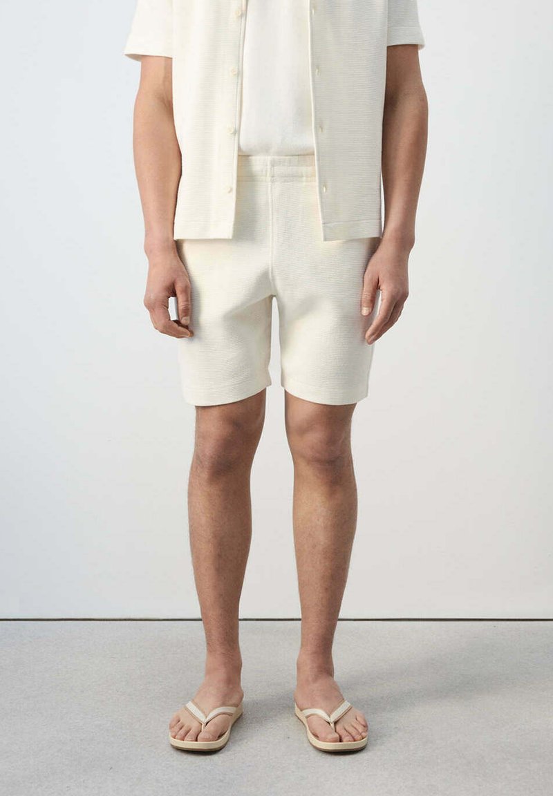 Modèle homme portant un short en maille crème, une chemise boutonnée à manches courtes assortie et des tongs beige, debout sur un sol gris avec un fond blanc.