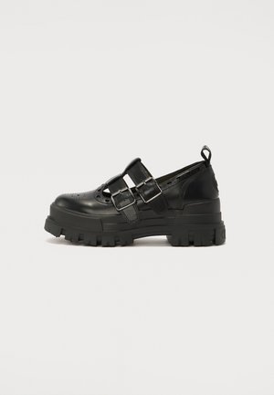 Black leather shoe with a rounded toe, featuring two adjustable buckles and perforated detailing, set on a thick, rugged sole.