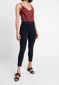 Röd leopardmönstrad bodysuit, högmidjade mörka jeans och svarta klacksandaler, framhävt mot en neutral bakgrund.