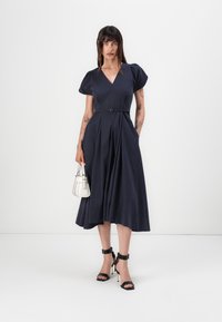Navyblaues Kleid mit Puffärmeln, V-Ausschnitt, tailliertem Gürtel und gefaltetem Rock. Kombiniert mit schwarzen High-Heel-Sandalen und einer kleinen weißen Handtasche.