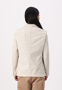 Light gray blazer, textured fabric, tailored fit, notched lapels, and two button accents on sleeves, worn over tan trousers.