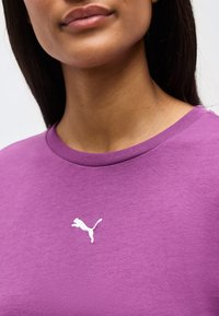 Νεαρή γυναίκα που φοράει μοβ μπλούζα με στρογγυλή λαιμόκοψη και μικρό λευκό λογότυπο Puma στο κέντρο του στήθους.