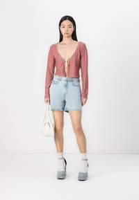 Haut à manches longues à carreaux rouges et blancs avec un décolleté en V profond et un nœud à l'avant, associé à un short en denim bleu clair. Le mannequin tient un petit sac blanc.