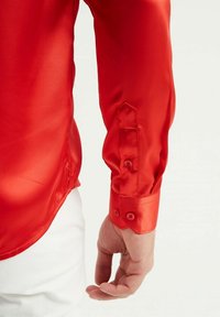 Chemise en satin rouge avec un col pointu, des poignets boutonnés et une texture lisse et brillante. Le design comprend une fente latérale à l'ourlet.