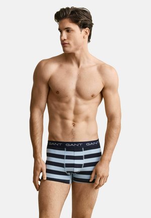 Homme torse nu aux cheveux courts et foncés, portant un boxer rayé bleu marine et bleu clair avec une ceinture "GANT", regardant vers sa gauche.