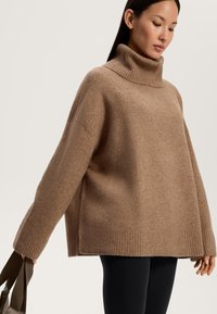 Maglione a collo alto in lana marrone lavorato a maglia con polsini e orlo a coste, caratterizzato da una vestibilità ampia e spacchi laterali. Abbinato a leggings neri.