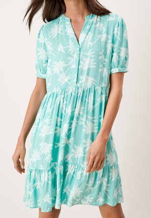 Jasnoturkusowa sukienka na krótki rękaw z białym nadrukiem palm, dekoltem w serek, marszczoną talią i falbaną u dołu, noszona przez stojącą kobietę.