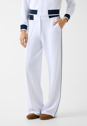 Femme portant un pantalon blanc texturé avec une ceinture bleu marine et des bandes latérales, associé à des chaussures plates marron, debout avec une main dans la poche.