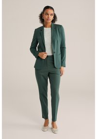 Temnozelena obleka z ozkimi hlačami in prilegajočim blaznikom, v kombinaciji s svetlo teksturirano bluzo in svetlo obutvijo s peto. Enostavni oblikovni detajli.