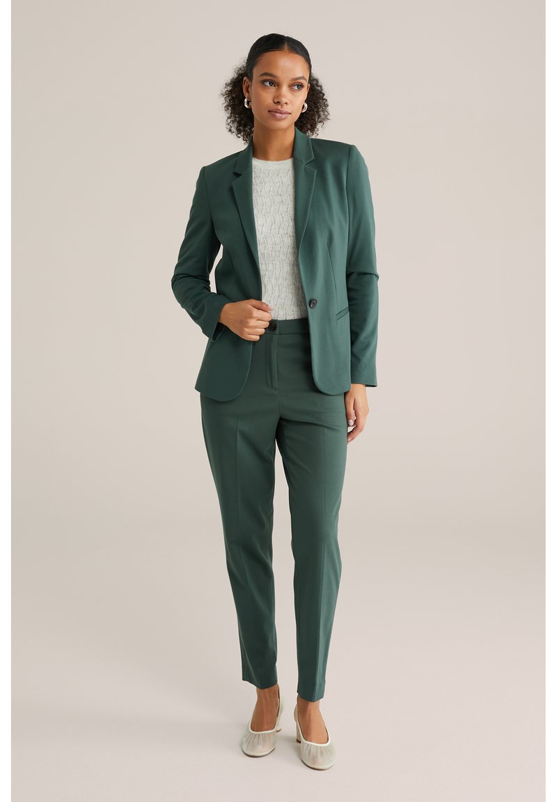 Temnozelena obleka z ozkimi hlačami in prilegajočim blaznikom, v kombinaciji s svetlo teksturirano bluzo in svetlo obutvijo s peto. Enostavni oblikovni detajli.