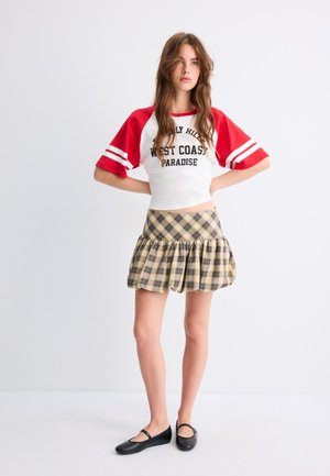 Jeune femme debout portant un haut rouge et blanc à manches courtes avec texte et une jupe courte beige à carreaux avec des ballerines noires sur fond blanc.