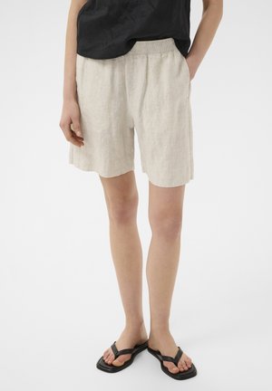 Persoon die beige linnen shorts, een zwarte mouwloze top en zwarte teenslippers met een vierkante neus draagt, staande op een witte achtergrond.