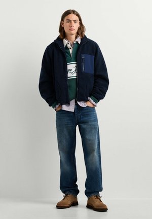 Giovane che indossa una giacca in pile blu navy, una camicia a righe verdi e bianche, jeans blu e scarpe marroni, in piedi con le mani nelle tasche.