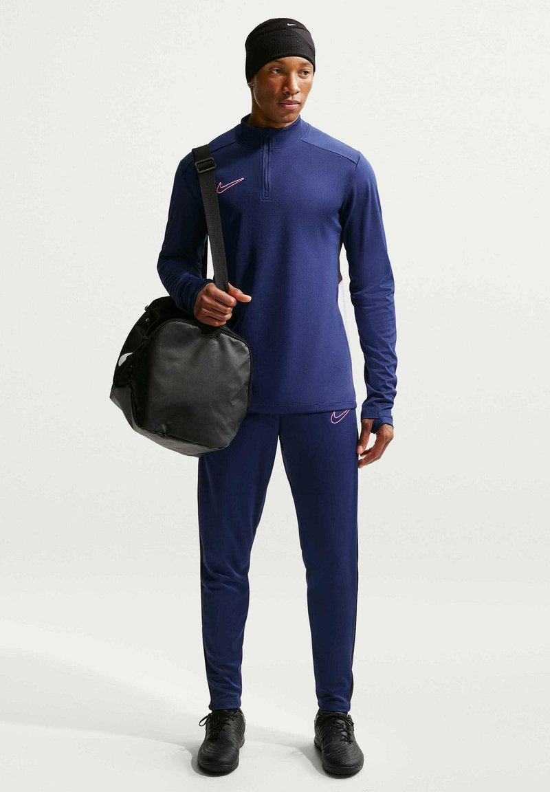 Mann trägt eine marineblaue Sport-Trainingsjacke und eine schwarze Mütze, hält eine schwarze Sporttasche über der Schulter und steht vor einem weißen Hintergrund.