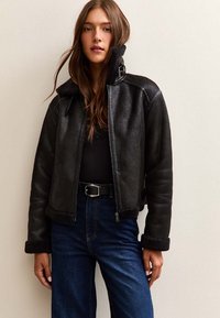 Giacca di pelle nera con colletto alto e chiusura a zip, con dettagli in shearling sui polsi e sull'orlo, abbinata a jeans in denim scuri.