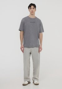 T-shirt in cotone grigio con una vestibilità rilassata, scollatura rotonda e dettaglio testuale "WEST COAST". Abbinato a jeans larghi grigio chiaro e sneakers.