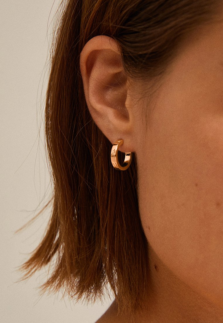 Pendiente en forma de aro de oro rosa, con forma redonda y acabado brillante. Textura suave y diseño minimalista visible contra el tono de piel y el cabello.