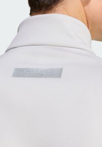 Lys grå pullover med høj krave. Har et sølvmærke på bagsiden med "SILVER ARROWS" præget. Glat tekstur og tætsiddende design.