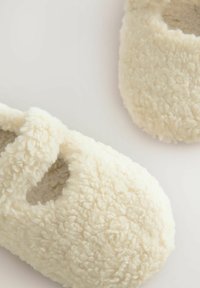 Krämfärgade tofflor gjorda av fluffigt, texturerat material, med en rundad form och en utskärning på ovansidan för andningsförmåga.