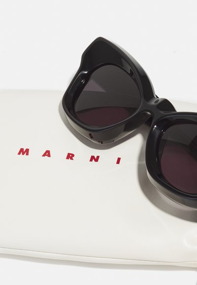 Occhiali da sole neri a forma di gatto con lenti scure, poggiati su una custodia bianca con il testo rosso in grassetto "MARNI". Superficie liscia e lucida.
