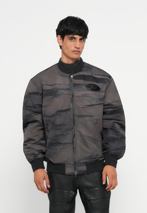 Gray bomber jacket with a textured, camo pattern, zip closure, ribbed cuffs, and a logo patch on the chest. Worn over black pants.