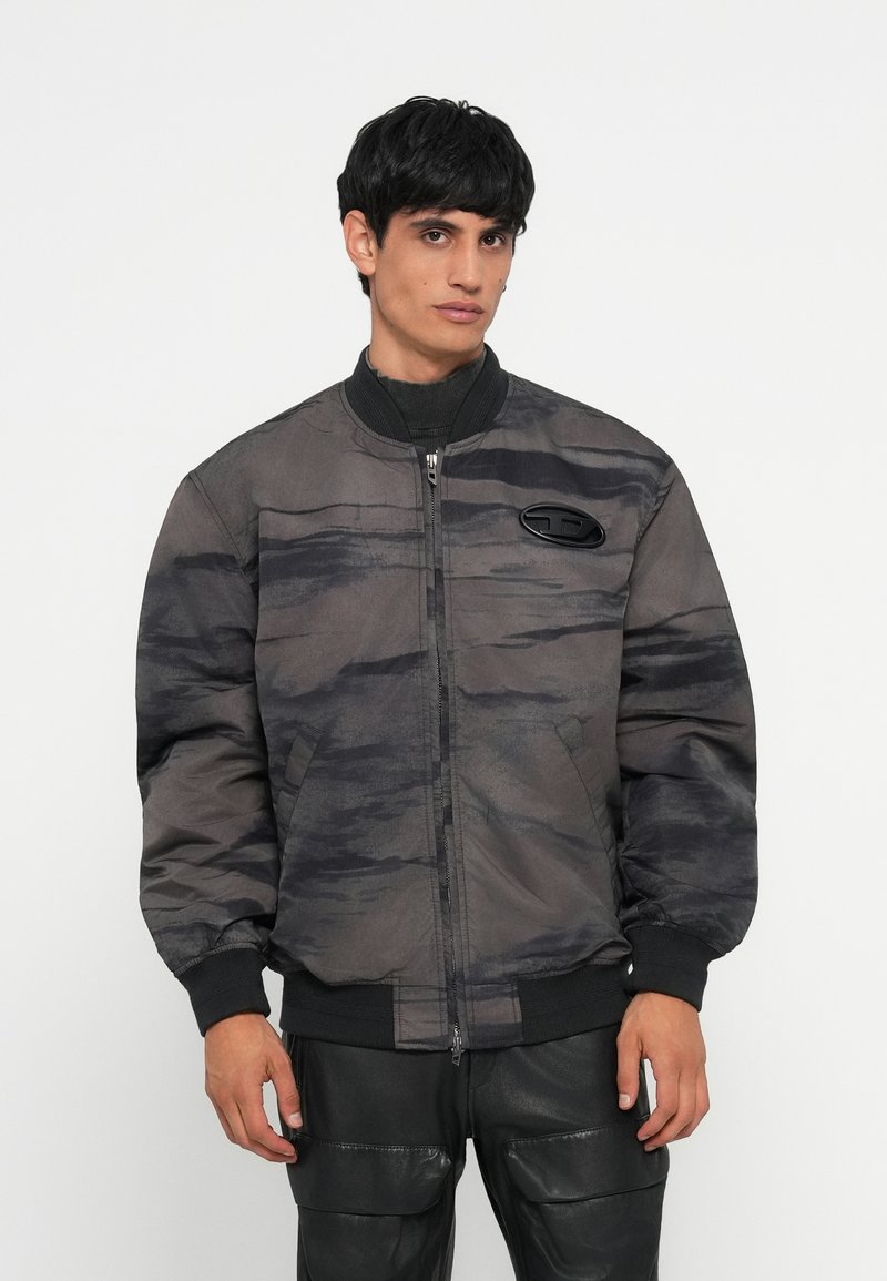Siv bomber jakna z teksturiranim kamuflažnim vzorcem, z zapenjanjem na zadrgo, rebrastimi manšetami in logotipom na prsih. Obuta nad črnimi hlačami.