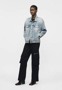 Femeie purtând o geacă largă de blugi de culoare albastru deschis, pantaloni cargo negri cu buzunare mari și cizme negre cu toc, stând în picioare pe fundal alb.