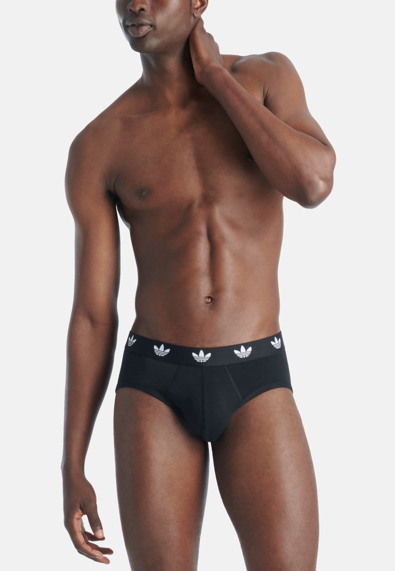 Homme torse nu portant un slip noir avec des logos tréfle blancs sur la ceinture, debout devant un fond clair uni.