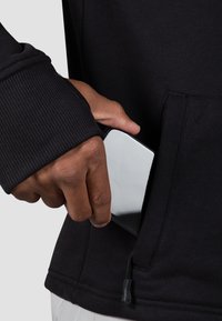 Čierna mikina s rebrovanými manžetami, obsahuje prednú kapsu. Ruka vkladá smartfón do vrecka, pričom predvádza hladký kovový okraj.