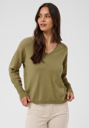 Frau mit langen dunklen Haaren, die einen olivgrünen V-Ausschnittpullover und hellbeige Hose trägt, schaut leicht zur Seite und hat die Hände nahe am Saum.