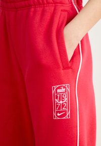 Punased dressipüksid, mis on valmistatud puuvilla segukangast, valgete külgdetailide ja trükitud logoga, millel on "Nike," "1972" ja lauri kujunduse aktsendid.