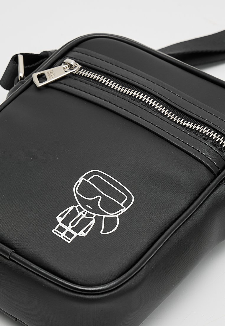 Bolso bandolera sintético negro con cierre superior de cremallera, con un gráfico de dibujos animados en blanco y herrajes de cremallera plateados. Textura suave.