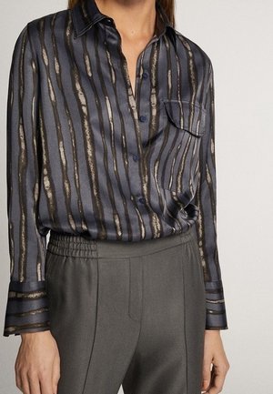 Woman wearing a silk button-up shirt with vertical stripes in black, brown, and beige, paired with high-waisted gray trousers.