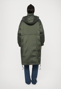 Long manteau de doudoune vert olive avec une capuche, cordon de serrage élastique à l'ourlet et manches larges. Pantalon en jean bleu évasé porté en dessous.