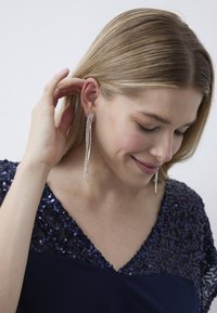 Orecchini pendenti con tonalità argento, caratterizzati da numerosi cristalli trasparenti disposti in fila, creando un effetto a cascata. Indossati da una donna.