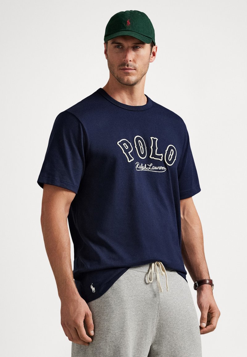 Moški z temno zeleno kapo, mornarsko modro majico "POLO Ralph Lauren", svetlo sivimi trenirkami in uro, stoji ob belem ozadju.