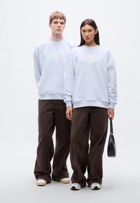 Ανοιχτό μπλε φούτερ με λευκά λογότυπα, συνδυασμένα με σκούρο καφέ φαρδιά παντελόνια. Το αρσενικό μοντέλο φοράει κρεμ παπούτσια· το θηλυκό μοντέλο κρατάει μια μαύρη τσάντα.