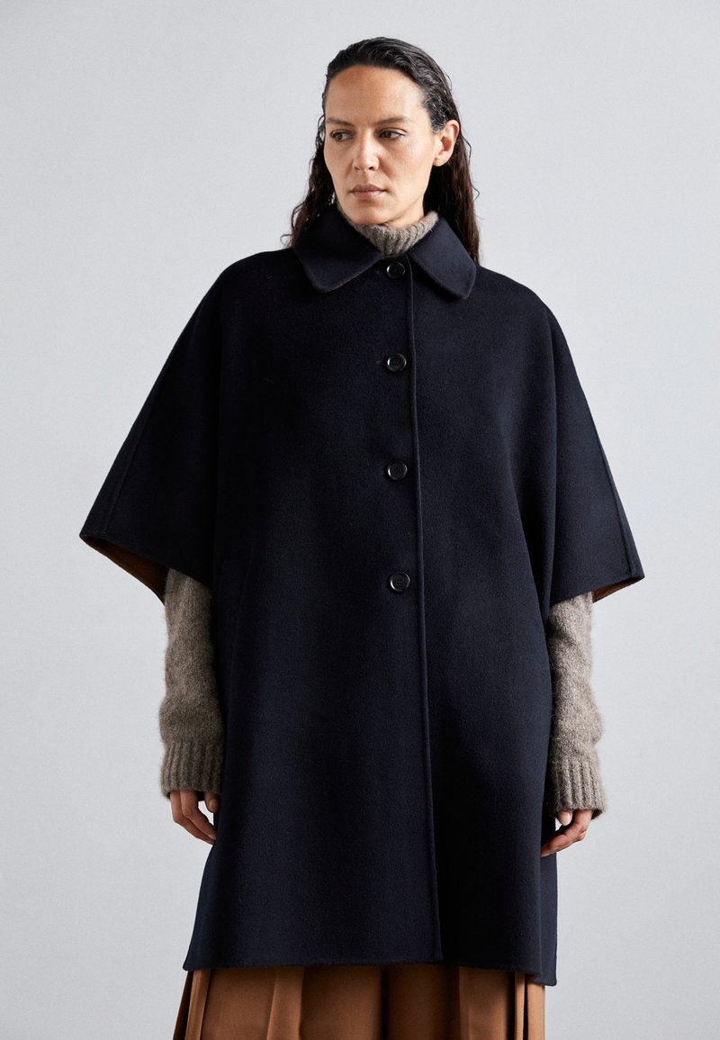 Cappotto a mantella in misto lana navy con collare a risvolto, cinque bottoni e maniche oversize, abbinato a un maglione beige in maglia sotto.