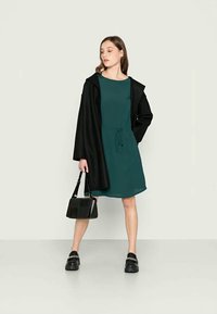 Vestido verde com cintura ajustável, usado por baixo de um longo casaco preto. O modelo carrega uma pequena bolsa preta e usa sapatos pretos robustos.