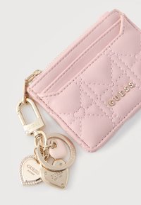 Ροζ ανοιχτόχρωμο καπιτονέ θήκη καρτών με χρυσά γράμματα "GUESS", συνδεδεμένο χρυσό μπρελόκ και καρδιές-μπρελόκ με ανάγλυφο κείμενο "GUESS EST. 1981."