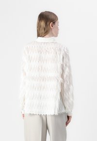 Blusa bianca trasparente con motivo a zigzag testurizzato, dotata di maniche lunghe, colletto e orlo inferiore sfrangiato. Tessuto leggero e arioso.